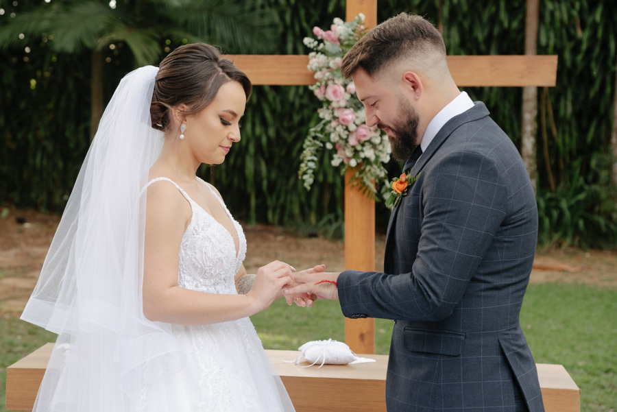 Noiva Thayná coloca a aliança no dedo do noivo Diego durante cerimônia ao ar livre no Espaço Verde Vale, em Taubaté. Fotografia de casamento com foco na troca de alianças em frente a uma cruz de madeira, capturada pelo fotógrafo Gilson Dias.