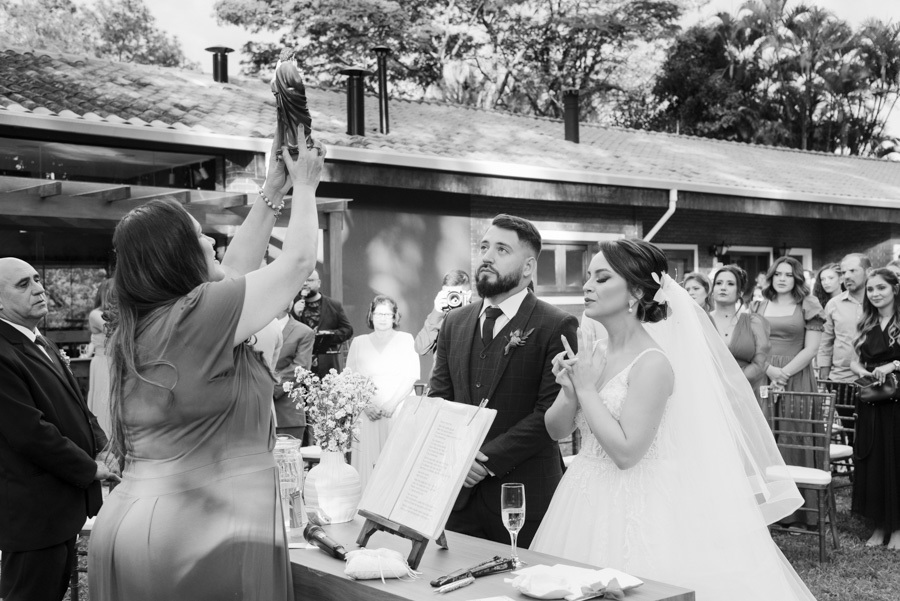 Fotografia em preto e branco de momento religioso na cerimônia de Thayná e Diego no Espaço Verde Vale, Taubaté. Celebrante levanta imagem sacra diante dos noivos no altar ao ar livre, em registro solene capturado pelo fotógrafo Gilson Dias.