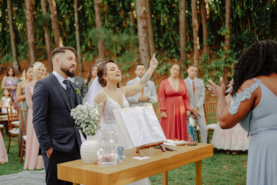 Noiva Thayná levanta a mão em momento de louvor e fé durante cerimônia ao ar livre no Espaço Verde Vale, Taubaté. Fotografia de casamento emocionante destacando a espiritualidade do casal, registrada pelo fotógrafo Gilson Dias no Vale do Paraíba.