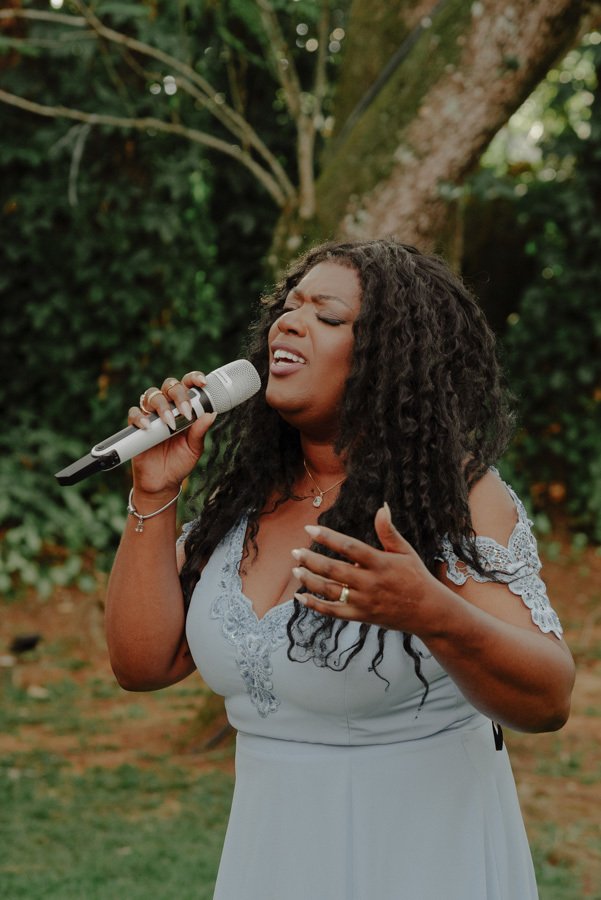 Cecília Militão com vestido azul claro canta emocionada durante cerimônia de casamento ao ar livre no Espaço Verde Vale, em Taubaté. Registro espontâneo de momento musical capturado pelo fotógrafo Gilson Dias no Vale do Paraíba.