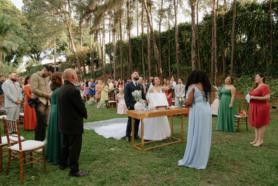 Noivos Thayná e Diego no altar durante momento de louvor em cerimônia ao ar livre no Espaço Verde Vale, Taubaté. Cena ampla com convidados, madrinhas e cantora solista sob as árvores, capturada pelo fotógrafo Gilson Dias no Vale do Paraíba.