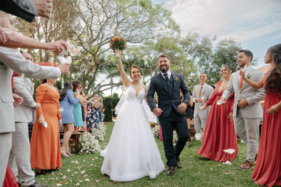 Noivos Thayná e Diego saem da cerimônia sob chuva de pétalas no Espaço Verde Vale, em Taubaté. Noiva levanta o buquê vibrante em comemoração, cercada por padrinhos em trajes terracota e cinza, em registro alegre por Gilson Dias Fotografia.