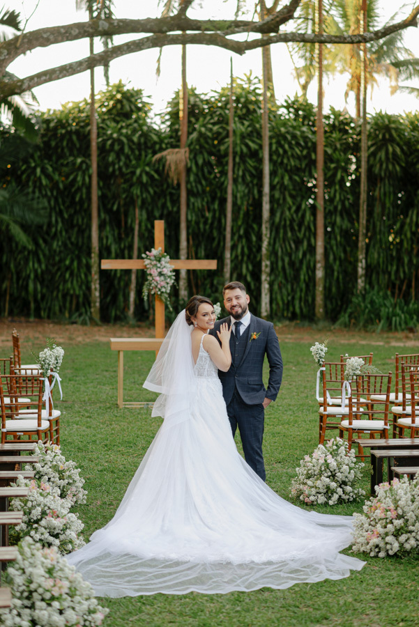 Noivos Thayná e Diego posam sorridentes no corredor da cerimônia ao ar livre no Espaço Verde Vale, Taubaté. Foto destaca a cauda do vestido da noiva e o altar com cruz de madeira ao fundo, registrada por Gilson Dias Fotografia no Vale do Paraíba.