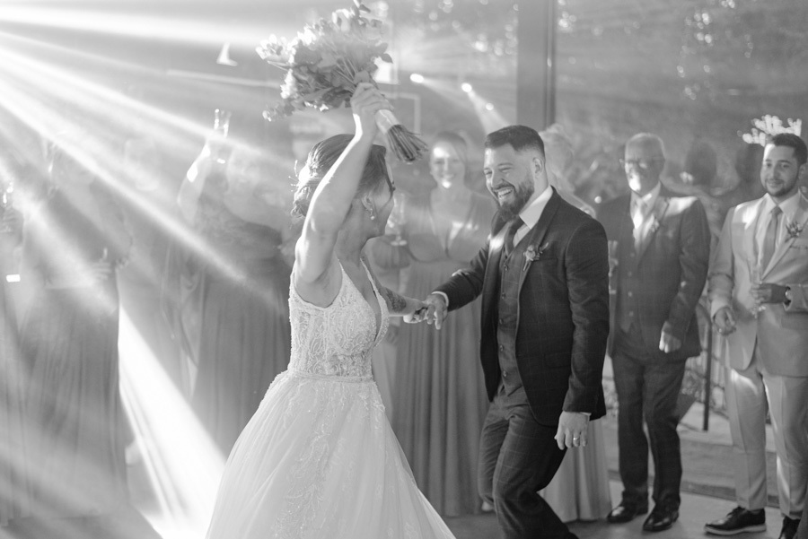 Noivos Thayná e Diego dançam animados na pista de dança do Espaço Verde Vale, em Taubaté. Fotografia em preto e branco capturando a noiva vibrando com o buquê para o alto sob feixes de luz, registrada por Gilson Dias Fotografia no Vale do Paraíba.