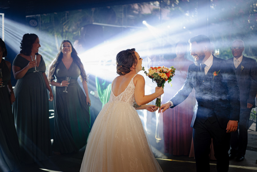 Noivos Thayná e Diego celebram na pista de dança do Espaço Verde Vale, em Taubaté. A noiva segura o buquê enquanto sorri para o noivo sob feixes de luz intensos e fumaça, em um registro vibrante e alegre por Gilson Dias Fotografia no Vale do Paraíba.