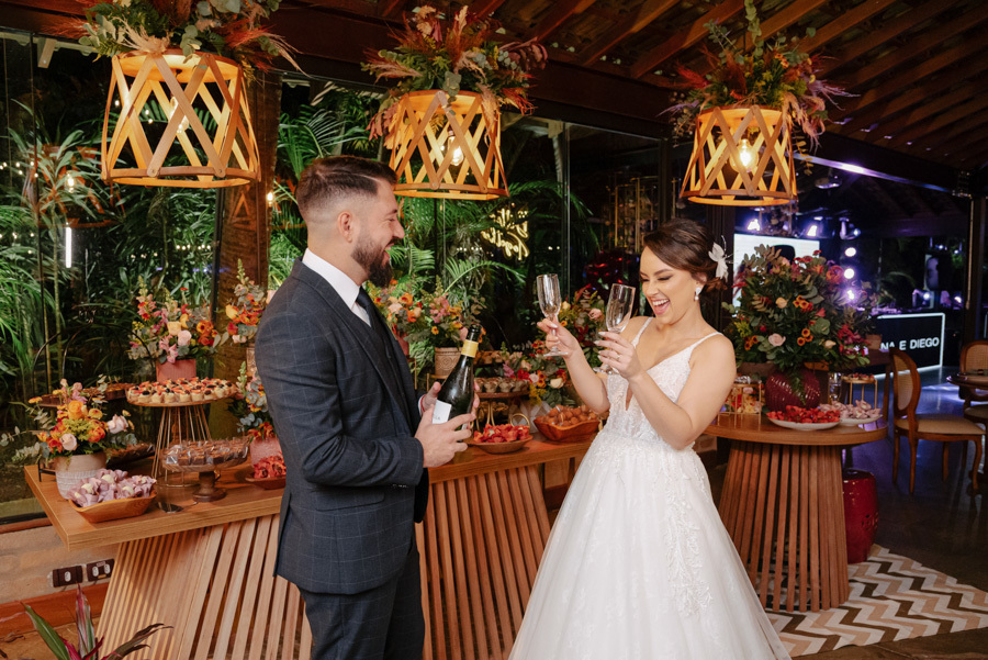 Noivos Thayná e Diego sorriem e brindam com espumante em frente à mesa de doces no Espaço Verde Vale, Taubaté. Fotografia de casamento destacando a decoração rústica, luminárias de palha e a alegria da recepção, registrada por Gilson Dias Fotografia.