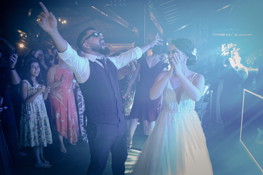 Noivos Thayná e Diego se divertem na pista de dança no Espaço Verde Vale, Taubaté. O noivo usa óculos escuros e vibra com os braços abertos enquanto a noiva aplaude sorridente sob luzes azuis, em registro animado por Gilson Dias Fotografia.