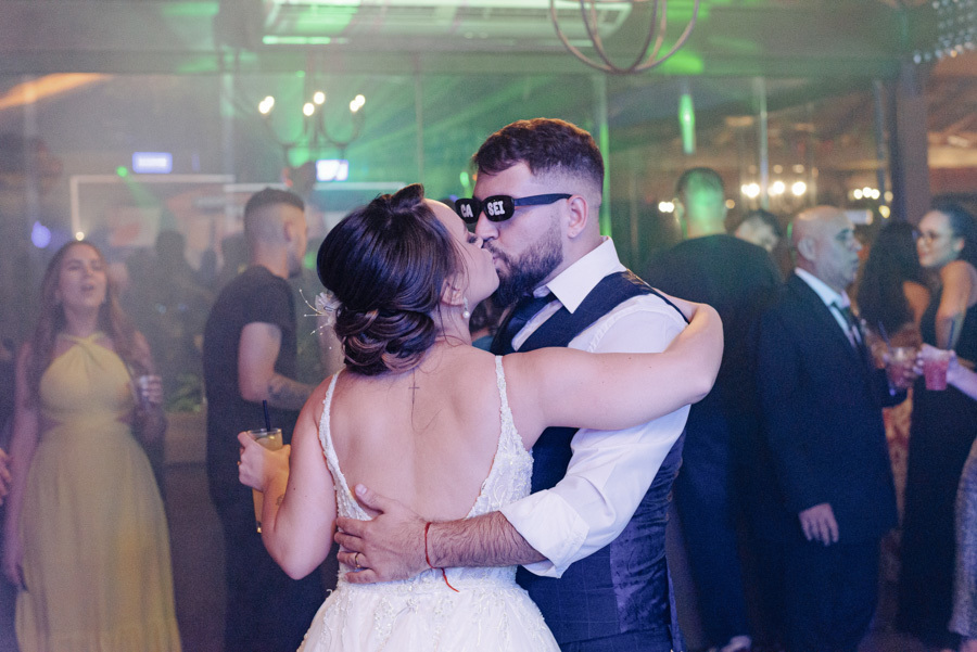 Noivos Thayná e Diego se beijam na pista de dança durante a festa no Espaço Verde Vale, Taubaté. Diego usa óculos divertidos com a frase 