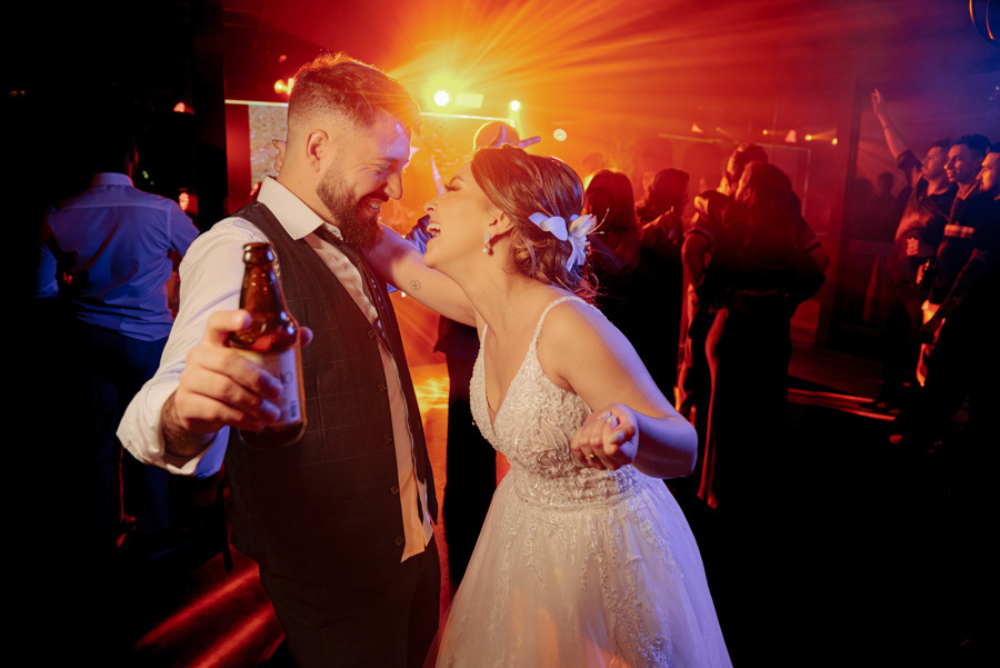 Noivos Thayná e Diego dão risada e dançam na pista do Espaço Verde Vale, Taubaté. Diego segura uma cerveja enquanto Thayná sorri sob luzes intensas em tons de vermelho e azul, em registro espontâneo da festa por Gilson Dias Fotografia.