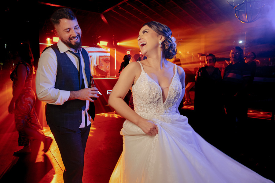Noiva Thayná gira seu vestido branco enquanto ri alegremente ao lado do noivo Diego na pista de dança. Fotografia de casamento vibrante com iluminação cênica vermelha e laranja no Espaço Verde Vale, Taubaté, registrada por Gilson Dias Fotografia.