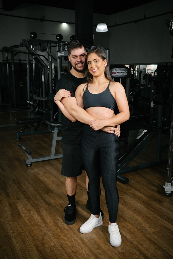 Matthaus e Rebeca sorriem abraçados em ensaio pré-wedding na Academia Aroeira. Registro espontâneo de fotógrafo profissional em Jacareí, capturando a cumplicidade e felicidade do casal em um ambiente que reflete sua rotina e paixão pela saúde.