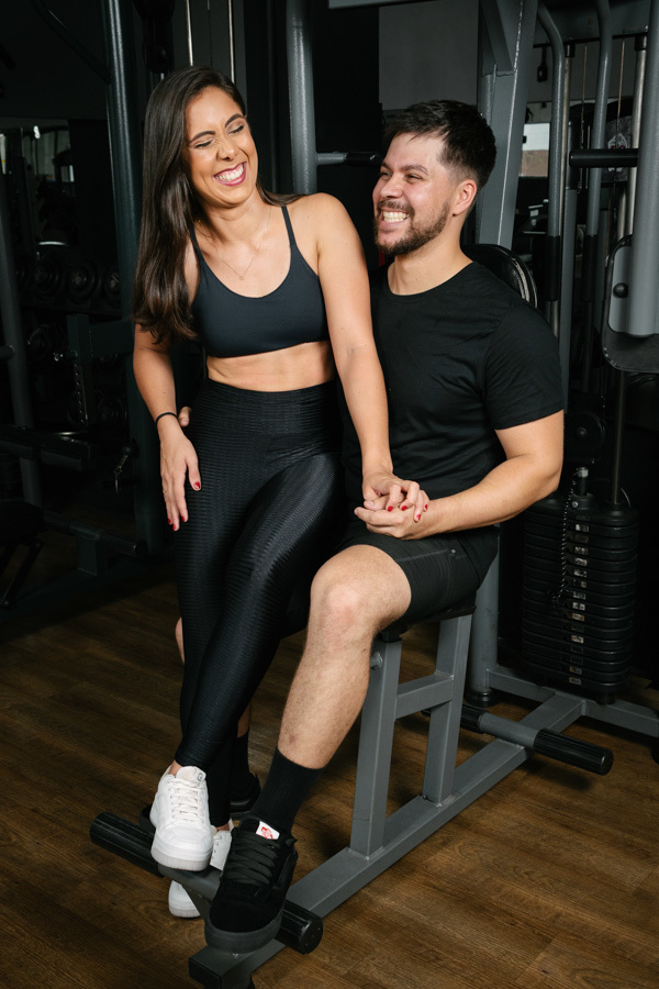 Matthaus e Rebeca dão risadas genuínas sentados em aparelho durante ensaio pré-wedding na Academia Aroeira. Foto de fotógrafo profissional em Jacareí, capturando a alegria e a leveza do casal em um registro espontâneo e cheio de vida.