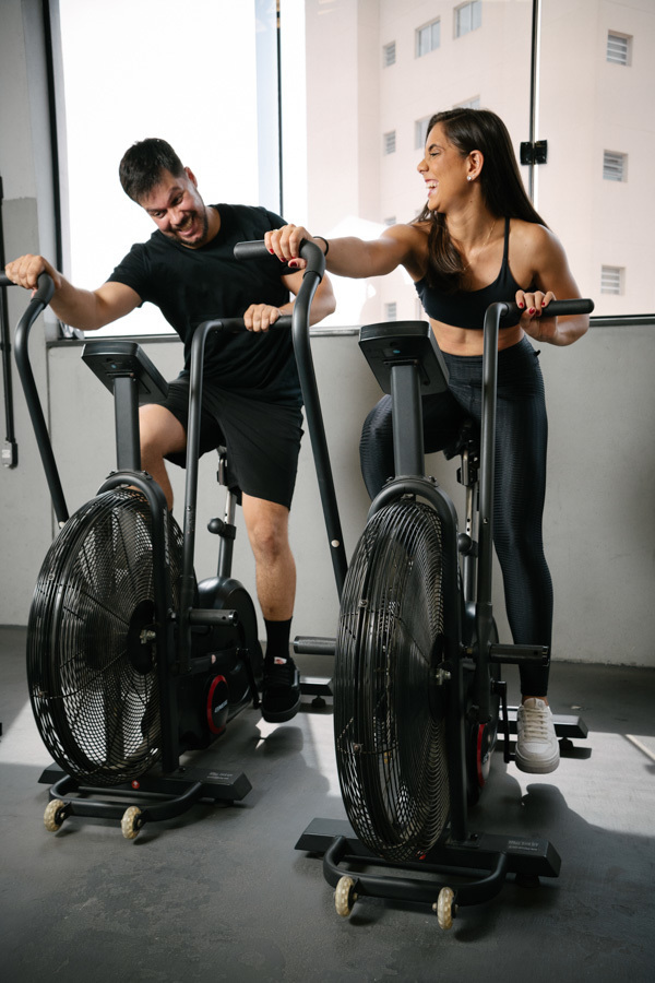 Matthaus e Rebeca se divertem pedalando em bicicletas ergométricas durante ensaio pré-wedding na Academia Aroeira. Foto espontânea de fotógrafo profissional em Jacareí, capturando a energia, alegria e parceria do casal em seu estilo de vida fitness.