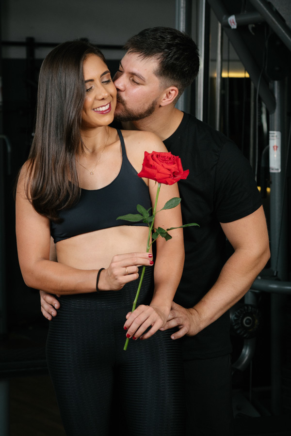 Matthaus beija o rosto de Rebeca, que segura uma rosa vermelha, em ensaio pré-wedding na Academia Aroeira. Foto de fotógrafo profissional em Jacareí unindo romantismo e o cenário fitness em um registro sensível e personalizado do casal.