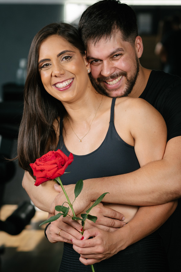 Matthaus e Rebeca sorriem abraçados com uma rosa vermelha em ensaio pré-wedding na Academia Aroeira. Foto de fotógrafo profissional em Jacareí, capturando o contraste entre o romantismo e o cenário fitness de forma espontânea e autêntica.