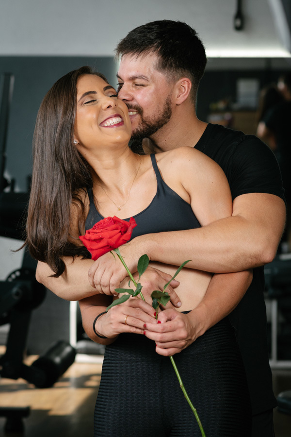Matthaus abraça e beija o rosto de Rebeca, que sorri segurando uma rosa vermelha, em ensaio pré-wedding na Academia Aroeira. Foto de fotógrafo profissional em Jacareí, capturando o romantismo e a cumplicidade do casal em um cenário fitness autêntico.