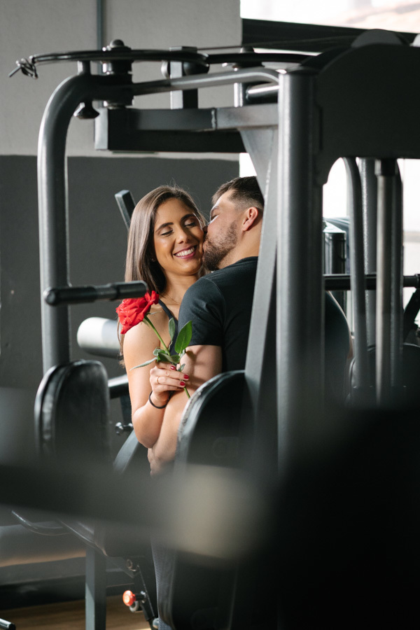 Matthaus beija o rosto de Rebeca, que sorri segurando uma rosa, em registro feito através de aparelhos de musculação. Foto de ensaio pré-wedding por fotógrafo profissional em Jacareí, criando um olhar íntimo e criativo dentro da Academia Aroeira.