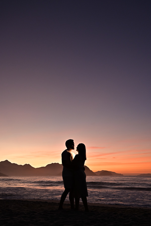 Silhueta do casal Isabela e Isaac no ensaio pré-wedding na Praia do Félix, Ubatuba. Fotógrafo profissional captura o casal abraçado sob o nascer do sol laranja e montanhas ao fundo, destacando o cenário natural e o clima romântico do litoral.