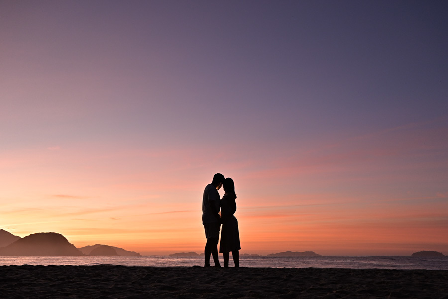 Fotografia de casal na Praia do Félix, Ubatuba. Isabela e Isaac em silhueta contra o horizonte do mar e céu colorido de manhã. Exemplo de ensaio fotográfico profissional com foco em espontaneidade, luz natural e a atmosfera única da praia.