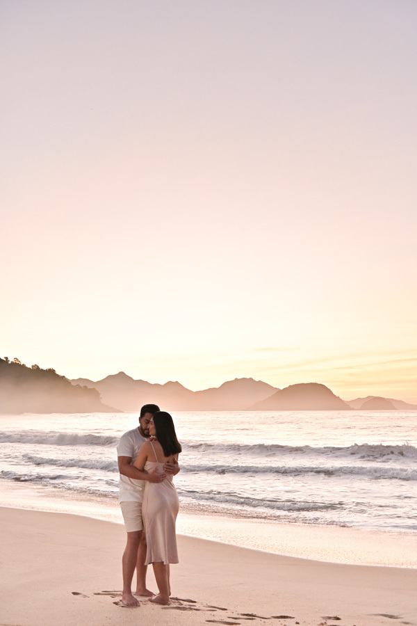 Isabela e Isaac abraçados na areia da Praia do Félix em Ubatuba. Foto com cores naturais e luz suave da manhã, mostrando os detalhes do casal e o mar ao fundo. Fotografia de casamento documental que captura conexão e leveza à beira-mar.