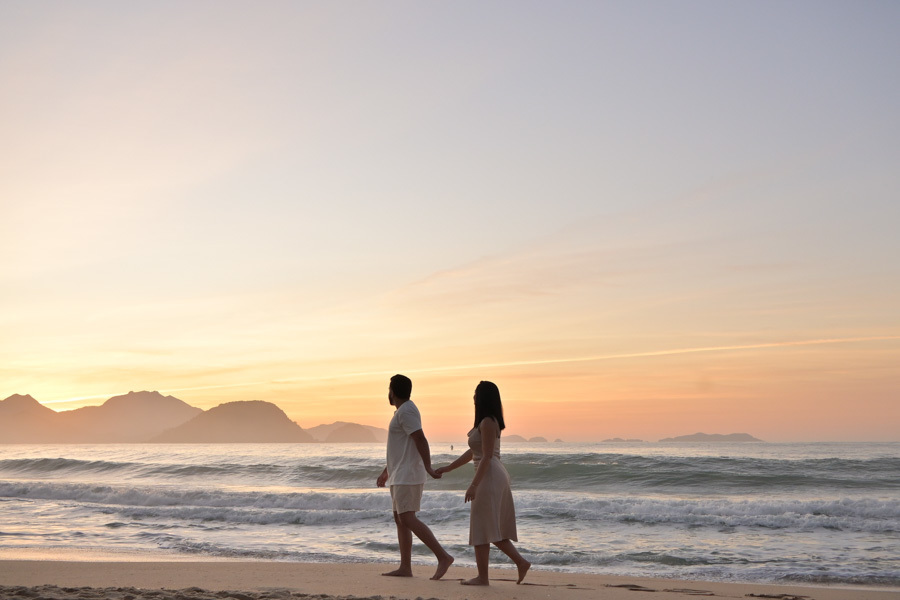 Silhueta de Isabela e Isaac caminhando de mãos dadas na beira do mar durante ensaio pré-wedding na Praia do Félix, Ubatuba. Fotografia documental com luz suave do amanhecer, destacando a conexão do casal e a paisagem litorânea de São Paulo.