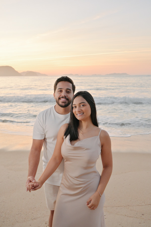 Retrato frontal de Isabela e Isaac sorrindo e de mãos dadas na areia da Praia do Félix, Ubatuba. Fotografia com cores naturais e luz clara da manhã, destacando a felicidade do casal e o mar ao fundo durante ensaio pré-casamento profissional.