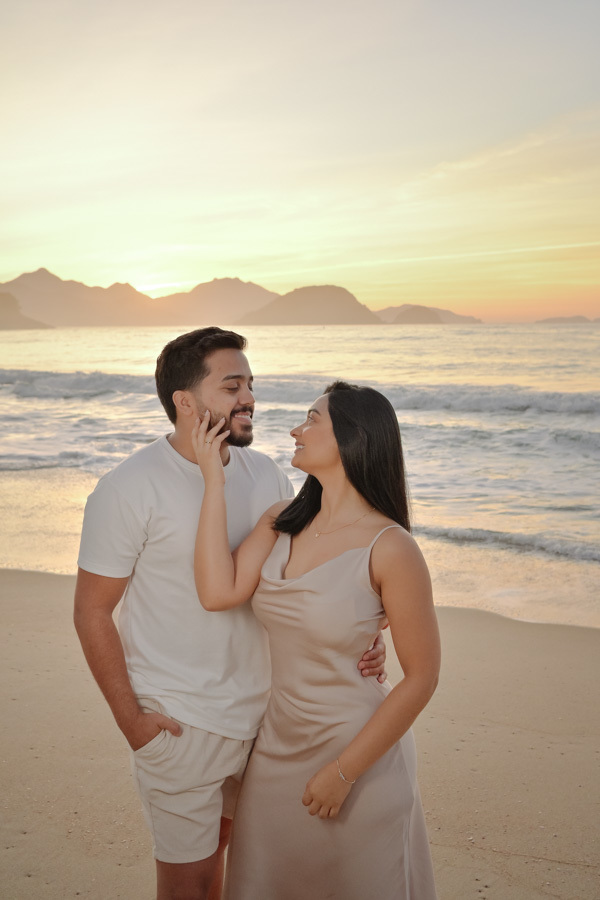 Isabela toca o rosto de Isaac em um momento de carinho na Praia do Félix, Ubatuba. Fotografia de ensaio pré-casamento com cores claras, luz suave do amanhecer e montanhas ao fundo, destacando a conexão e o romantismo do casal no litoral.