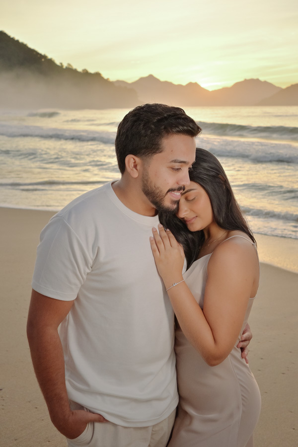 Isabela repousa a cabeça no peito de Isaac em um abraço acolhedor na Praia do Félix, Ubatuba. Fotografia de ensaio pré-casamento com cores claras, luz suave de amanhecer e névoa nas montanhas ao fundo, transmitindo paz e conexão profunda do casal.
