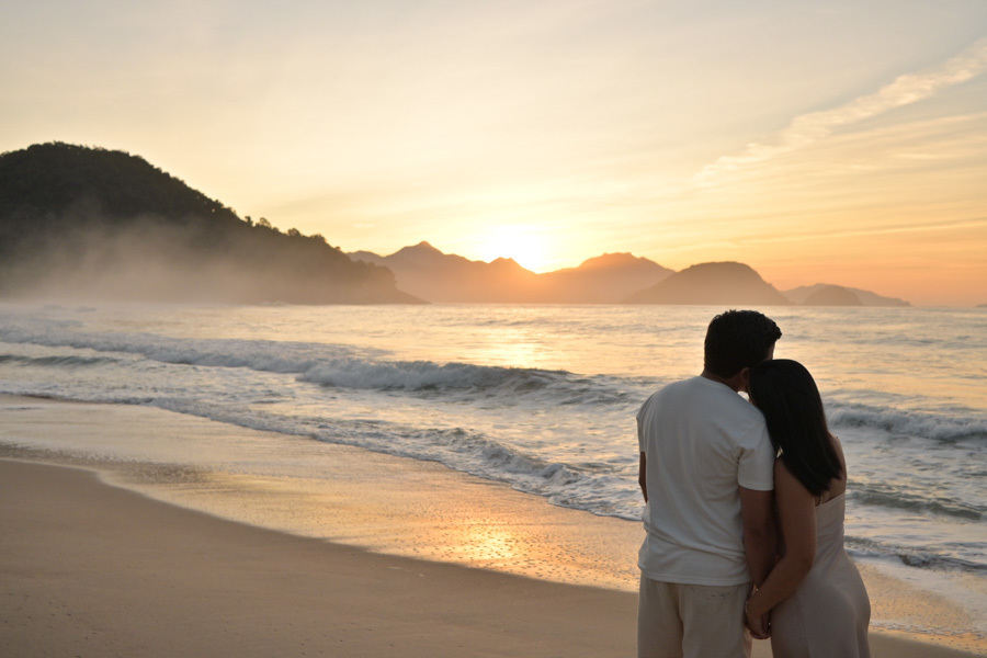 Casal Isabela e Isaac vistos de costas, abraçados e observando o nascer do sol na Praia do Félix, Ubatuba. Fotografia com luz dourada refletida no mar e névoa nas montanhas, encerrando o ensaio pré-wedding com um clima de paz e contemplação.