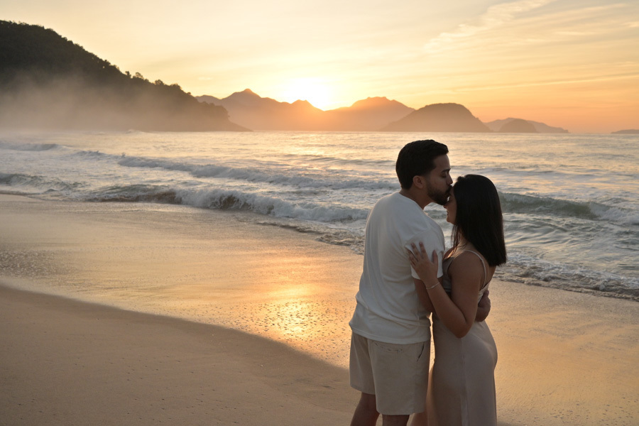 Isaac beija a testa de Isabela em um abraço carinhoso à beira-mar na Praia do Félix, Ubatuba. Fotografia de ensaio pré-wedding com luz dourada do sol refletida na água, montanhas ao fundo e uma atmosfera de amor e cumplicidade no litoral.