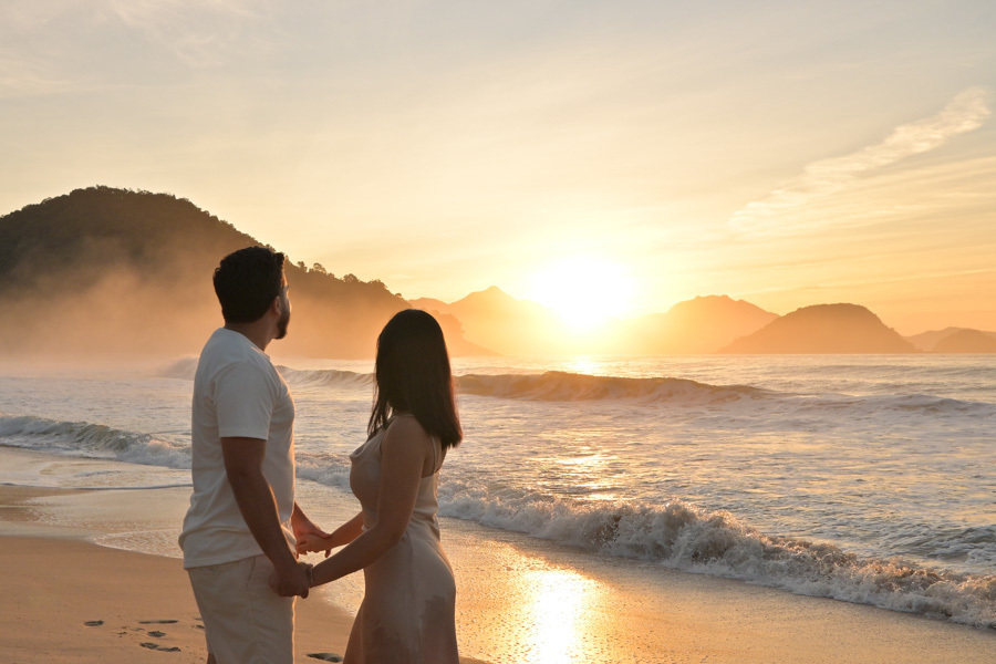 Isabela e Isaac de mãos dadas à beira-mar na Praia do Félix, Ubatuba, observando o sol radiante no horizonte. Fotografia profissional com luz intensa do amanhecer, névoa sobre as montanhas e reflexos dourados na areia molhada e nas ondas.