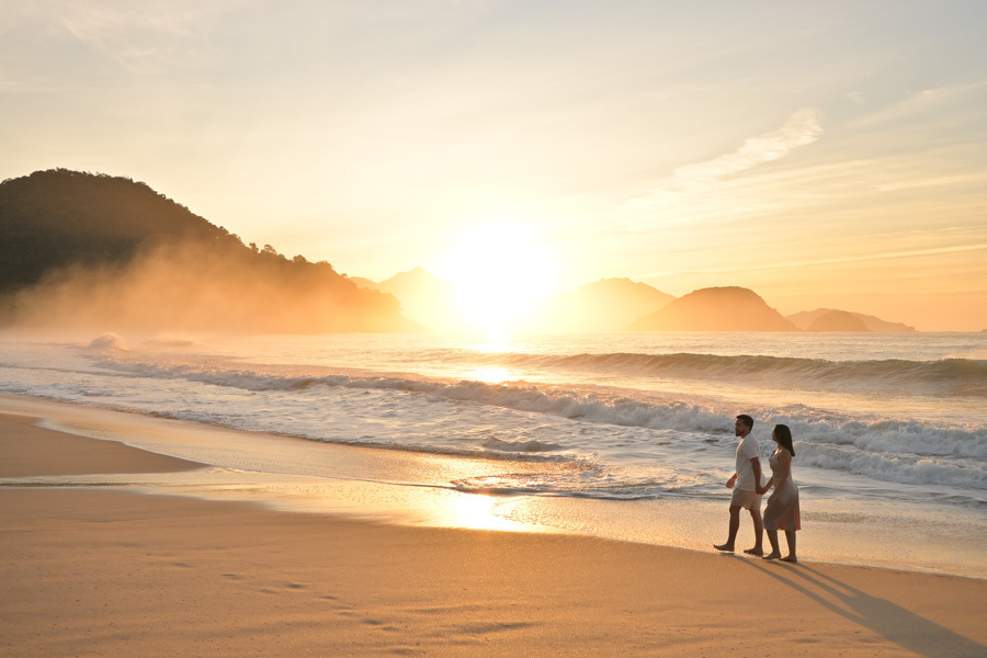 Isabela e Isaac caminham de mãos dadas pela areia molhada da Praia do Félix, em Ubatuba, sob um sol radiante. Fotografia profissional com luz de amanhecer intensa, névoa nas montanhas e reflexos dourados que destacam a vastidão e beleza do cenário.