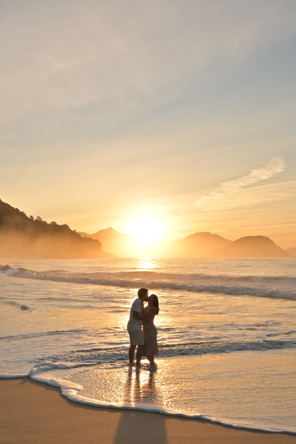Isabela e Isaac se beijam com os pés na água durante o amanhecer na Praia do Félix, Ubatuba. Fotografia profissional com sol radiante ao fundo, reflexos dourados nas ondas e névoa nas montanhas, capturando um momento mágico de amor no litoral norte.