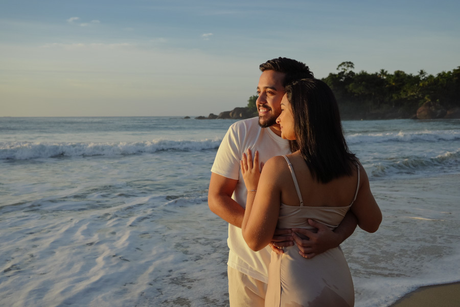 Isabela e Isaac abraçados na areia da Praia do Félix, em Ubatuba, observando o mar sob a luz suave da manhã. Fotografia profissional de pré-wedding com foco na expressão de felicidade do casal e na beleza natural do litoral norte de São Paulo.