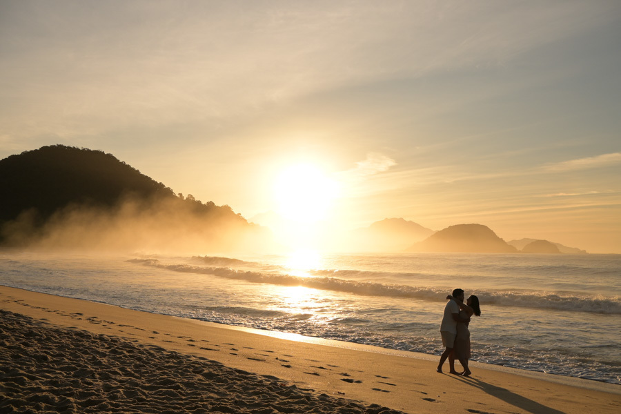 Isabela e Isaac se abraçam na areia da Praia do Félix, Ubatuba, sob a luz dourada de um sol radiante. Fotografia profissional de pré-wedding com névoa nas montanhas e reflexos intensos na água, capturando um momento mágico e caloroso à beira-mar.