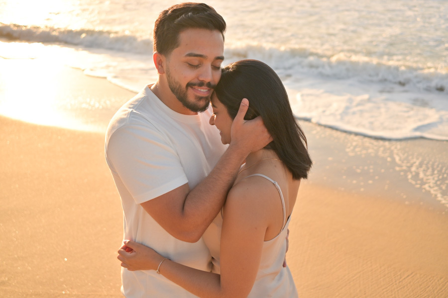 Close carinhoso de Isaac abraçando Isabela e tocando seu rosto suavemente na Praia do Félix, Ubatuba. Fotografia de pré-wedding com luz dourada do sol, cores quentes e o mar ao fundo, destacando a intimidade e a emoção do casal no litoral.