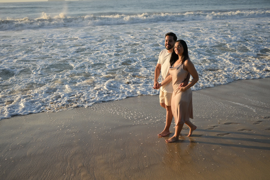 Isabela e Isaac caminham de mãos dadas pela areia molhada da Praia do Félix, em Ubatuba, com ondas suaves ao fundo. Fotografia profissional de pré-wedding com luz natural da manhã, capturando a alegria e o companheirismo do casal no litoral.