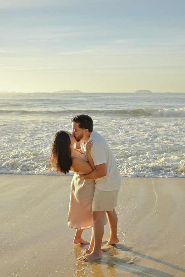 Isabela e Isaac se beijam apaixonadamente na beira do mar durante o ensaio pré-wedding na Praia do Félix, Ubatuba. Fotografia profissional com luz clara da manhã, ondas suaves e areia dourada, capturando um momento romântico e eterno no litoral.