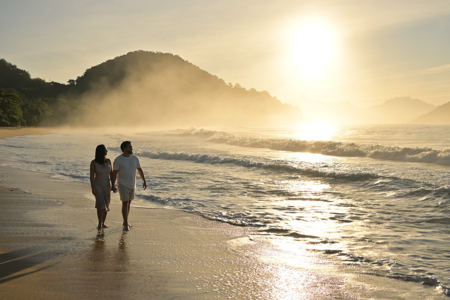 Isabela e Isaac caminham de mãos dadas pela areia dourada da Praia do Félix, em Ubatuba, durante o amanhecer. Fotografia profissional com luz solar intensa, névoa sobre a encosta e reflexos brilhantes nas ondas, destacando a beleza do litoral norte.