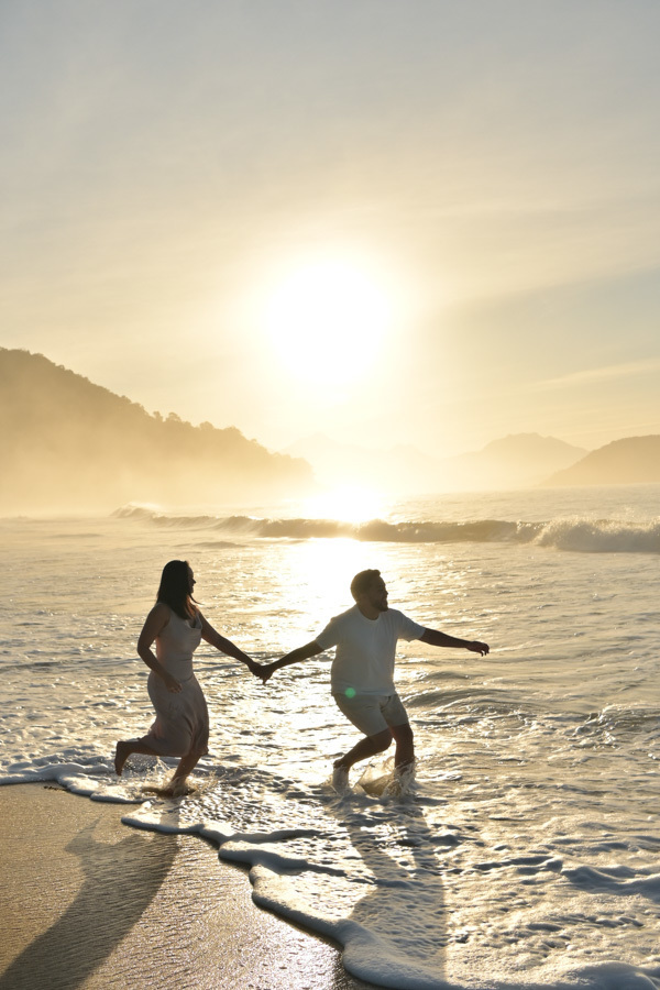 Isabela e Isaac correm de mãos dadas pela beira do mar na Praia do Félix, Ubatuba, durante um amanhecer radiante. Fotografia de pré-wedding cheia de energia, com luz solar intensa, reflexos na água e névoa sobre as montanhas ao fundo.