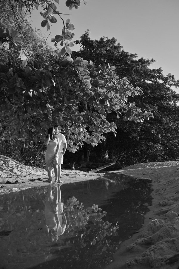 Isabela e Isaac em preto e branco na Praia do Félix, Ubatuba. O casal está sob árvores na areia, com seu reflexo nitidamente projetado em um pequeno curso d'água. Fotografia profissional com estilo artístico e documental no litoral de São Paulo.