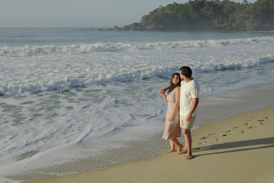 Isabela e Isaac caminham abraçados pela areia da Praia do Félix, em Ubatuba, com ondas brancas quebrando ao fundo. Fotografia profissional de pré-wedding com cores naturais, luz suave e foco na cumplicidade do casal no litoral norte de São Paulo.