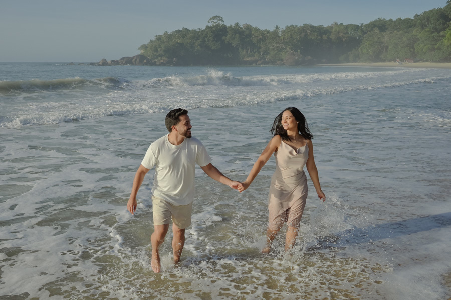 Isabela e Isaac correm sorridentes e de mãos dadas pela beira do mar na Praia do Félix, Ubatuba. Fotografia de pré-wedding com o casal se divertindo entre as ondas, sob luz natural clara, destacando a alegria e a natureza exuberante do litoral.