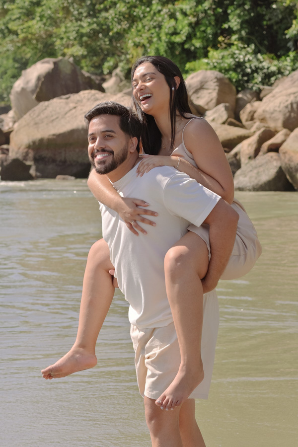 Isaac carrega Isabela nas costas (piggyback) enquanto caminham com os pés na água na Praia do Félix, Ubatuba. Fotografia de pré-wedding alegre e descontraída, com o casal sorrindo, pedras ao fundo e luz natural clara realçando a felicidade do momento.