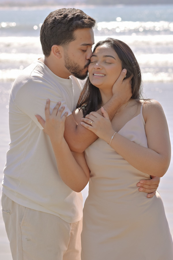 Isaac abraça Isabela por trás e beija seu rosto com carinho na Praia do Félix, Ubatuba. Fotografia de pré-wedding com luz natural clara, destacando a felicidade da noiva de olhos fechados e a cumplicidade do casal à beira-mar.