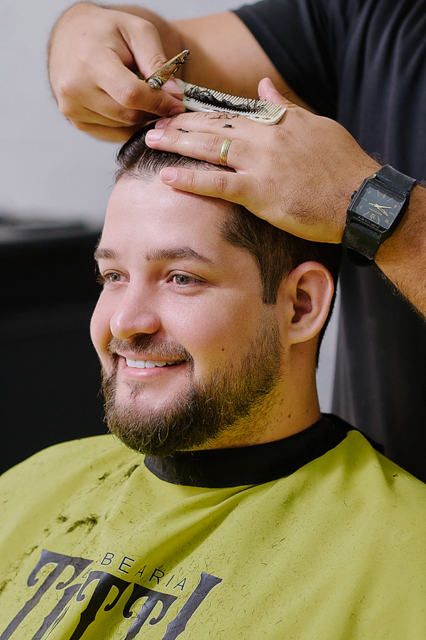Noivo Caciano sorridente durante o making of na Barbearia Totti em São José dos Campos. Fotografia documental de casamento capturando a preparação e alegria do noivo antes da cerimônia na Capela Nossa Senhora Aparecida.