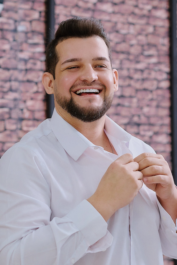 Noivo Caciano dando uma risada espontânea enquanto abotoa a camisa branca. Fotografia de casamento em São José dos Campos que traduz a alegria genuína e o clima descontraído do making of antes da cerimônia na Capela Nossa Senhora Aparecida.