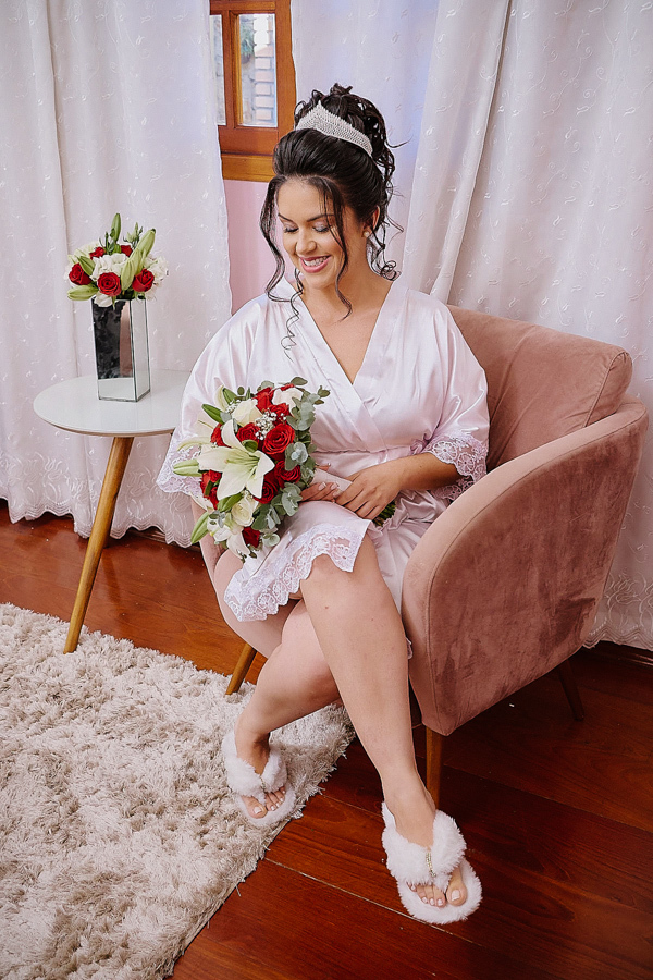Noiva Paloma sorridente sentada em uma poltrona, segurando seu buquê de rosas vermelhas e lírios brancos durante o making of em São José dos Campos. Fotografia de casamento capturando a alegria e os detalhes finais antes da cerimônia na Capela.