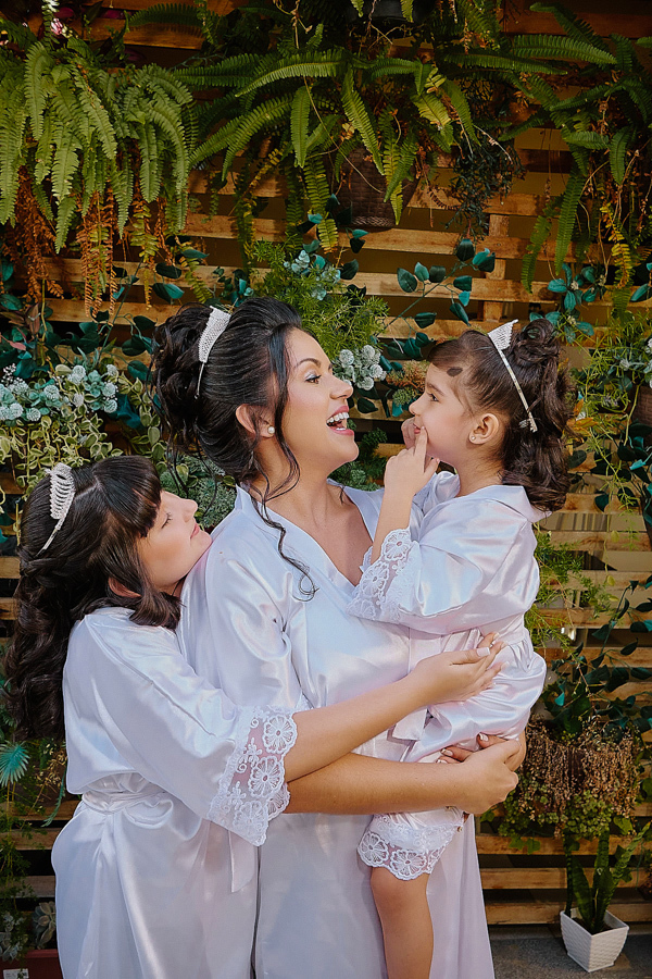 Noiva Paloma sorridente abraçada às suas duas filhas durante o making of em São José dos Campos. As três usam robas de cetim branco, celebrando a união da família em um momento de puro afeto antes da cerimônia na Capela Nossa Senhora Aparecida.