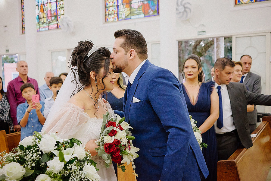 Noivo Cassiano beija a testa da noiva Paloma no altar da Capela Nossa Senhora Aparecida, em São José dos Campos. Um registro emocionante que captura a ternura e a solidez da união do casal diante de seus convidados e familiares.